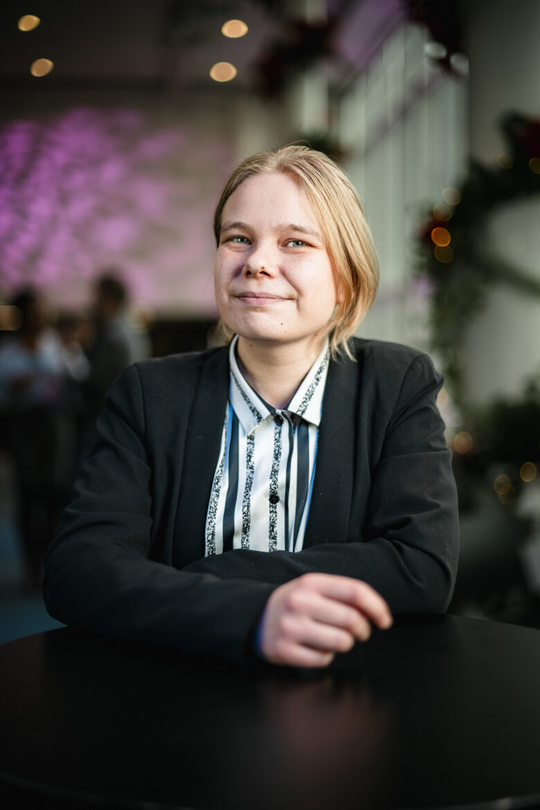 A young woman with blonde hair smiles at the camera, wearing a black blazer over a striped shirt. Her arms are crossed in front of her, resting on a dark table.