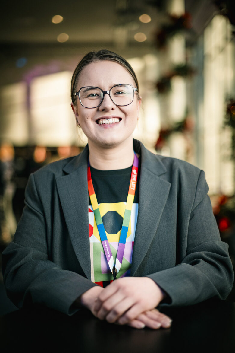 A young woman with glasses smiles at the camera. She wears a dark blazer over a black shirt with a colorful lanyard around her neck.